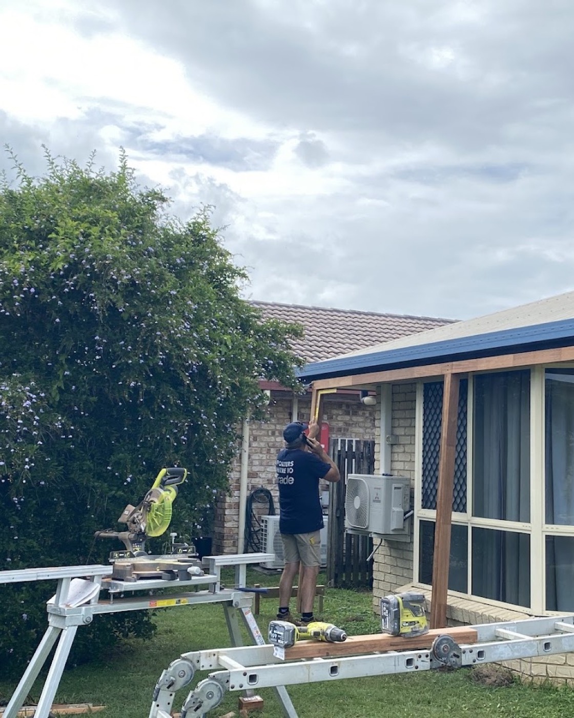 A tradesman on a step working on a new timber awning frame at a brick home, with a drop saw and trestles set up on the lawn