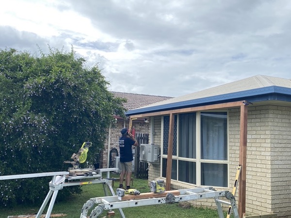 A tradesman on a step fixing a new timber awning frame to a brick home, with a drop saw and trestles on the lawn